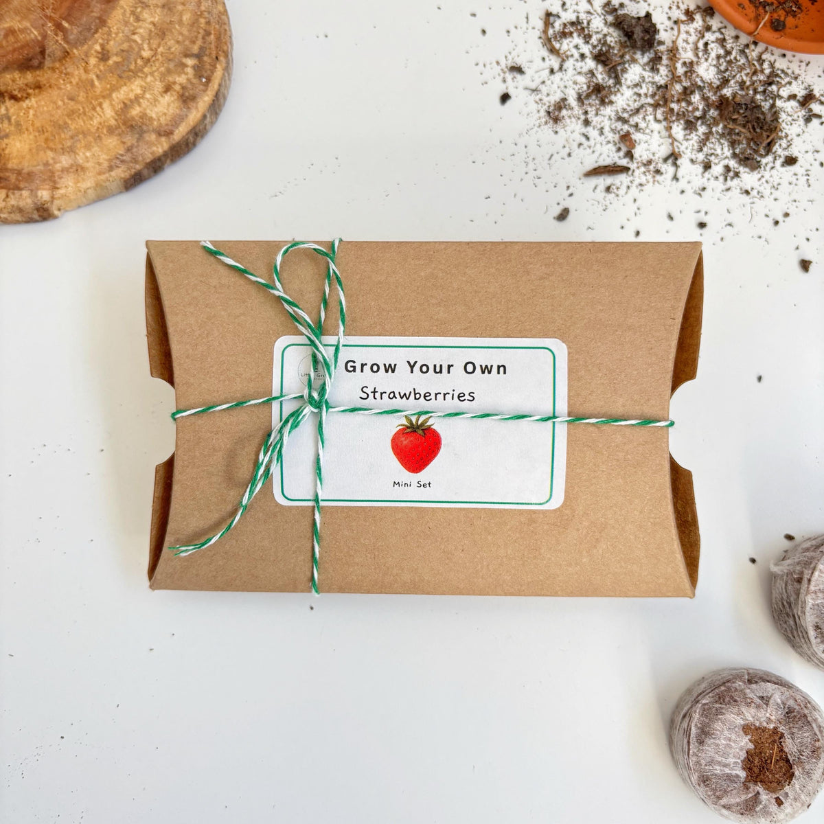 Kraft paper package with green string, 'Grow Your Own Strawberries' label, and small compost discs on a white surface.