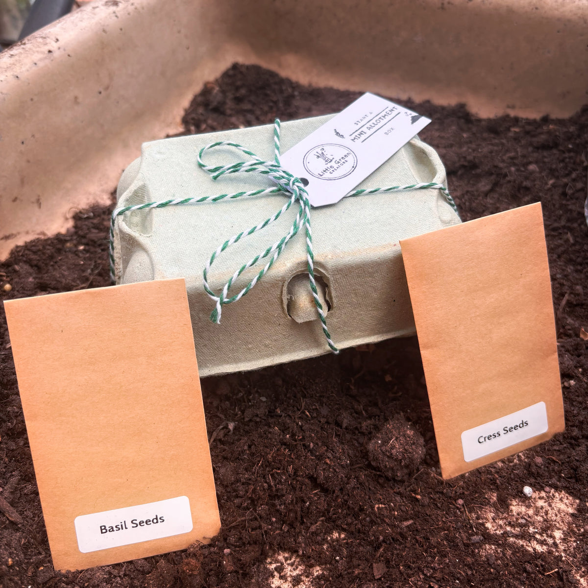 A small seed-starting packet wrapped in pale green paper and tied with green-and-white string sits in dark soil, with two brown paper seed labels in front reading 'Basil Seeds' and 'Cress Seeds'.