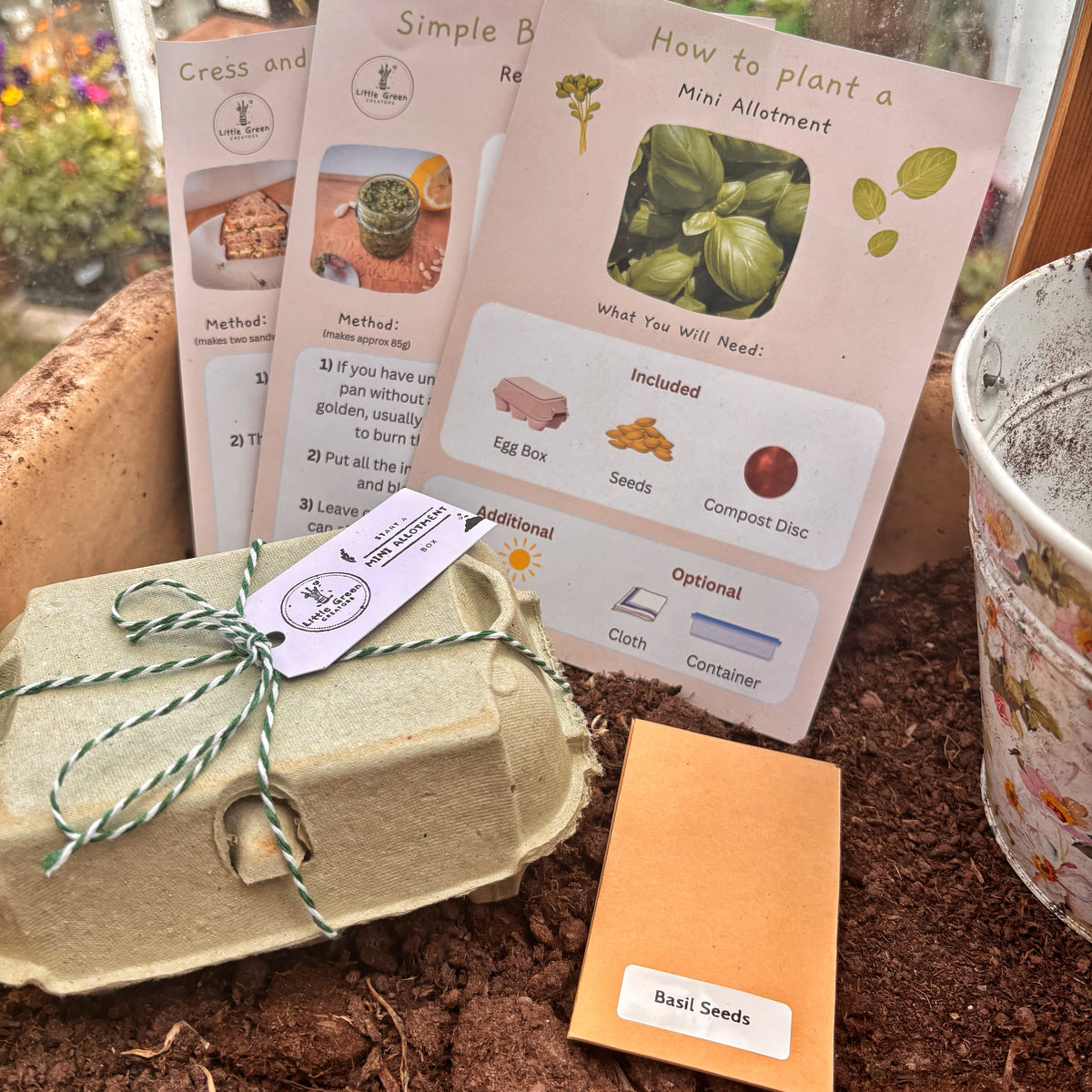 Start a Mini Allotment gardening starter kit components laid out on soil, including printed mini-allocation cards, a seed packet labeled Basil Seeds, a paper egg carton tied with string, and a floral pot.