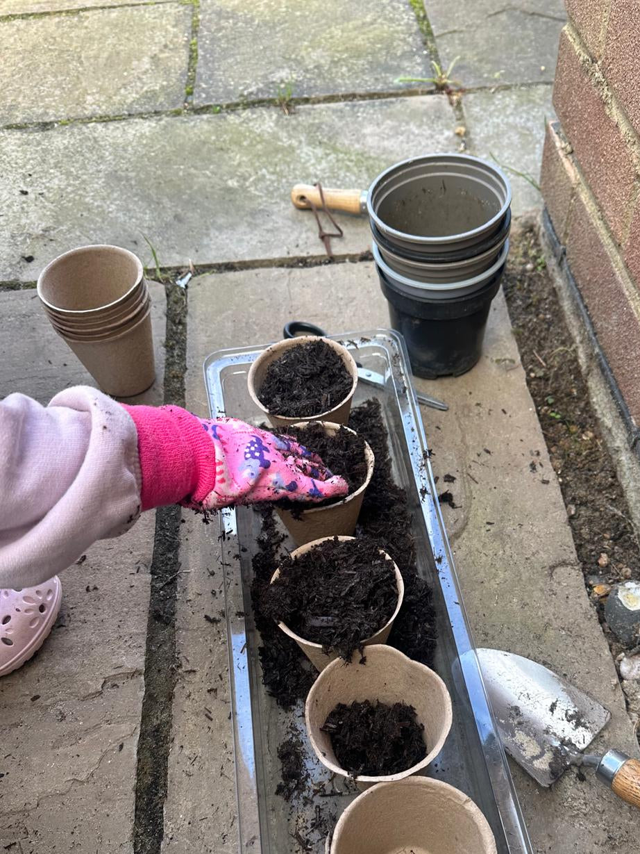 child with a pink glove putting hand in soil. Six biodegradable plant pots in a row on a tiled floor.